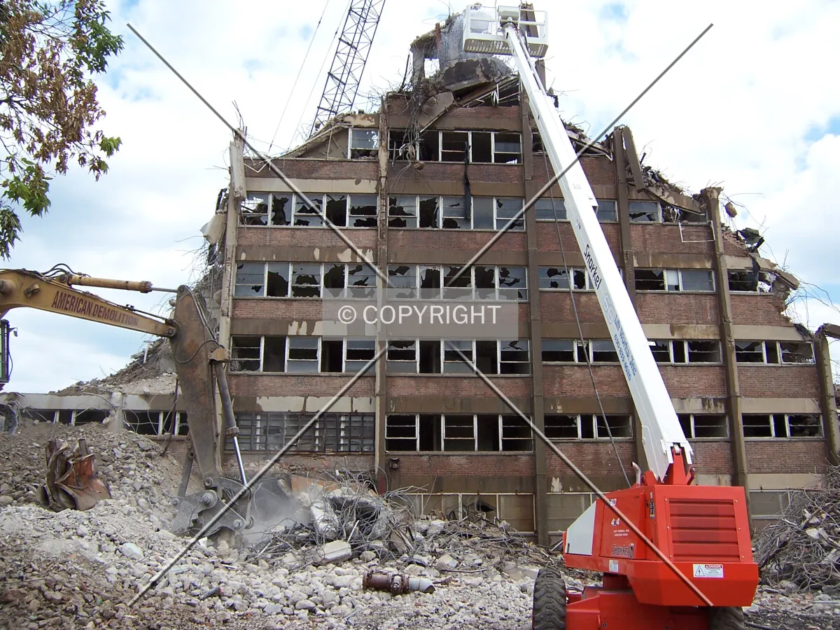 Demolition view from the west. A worker in the crane basket is spraying water on the structure to mitigate dust created by the demolition work