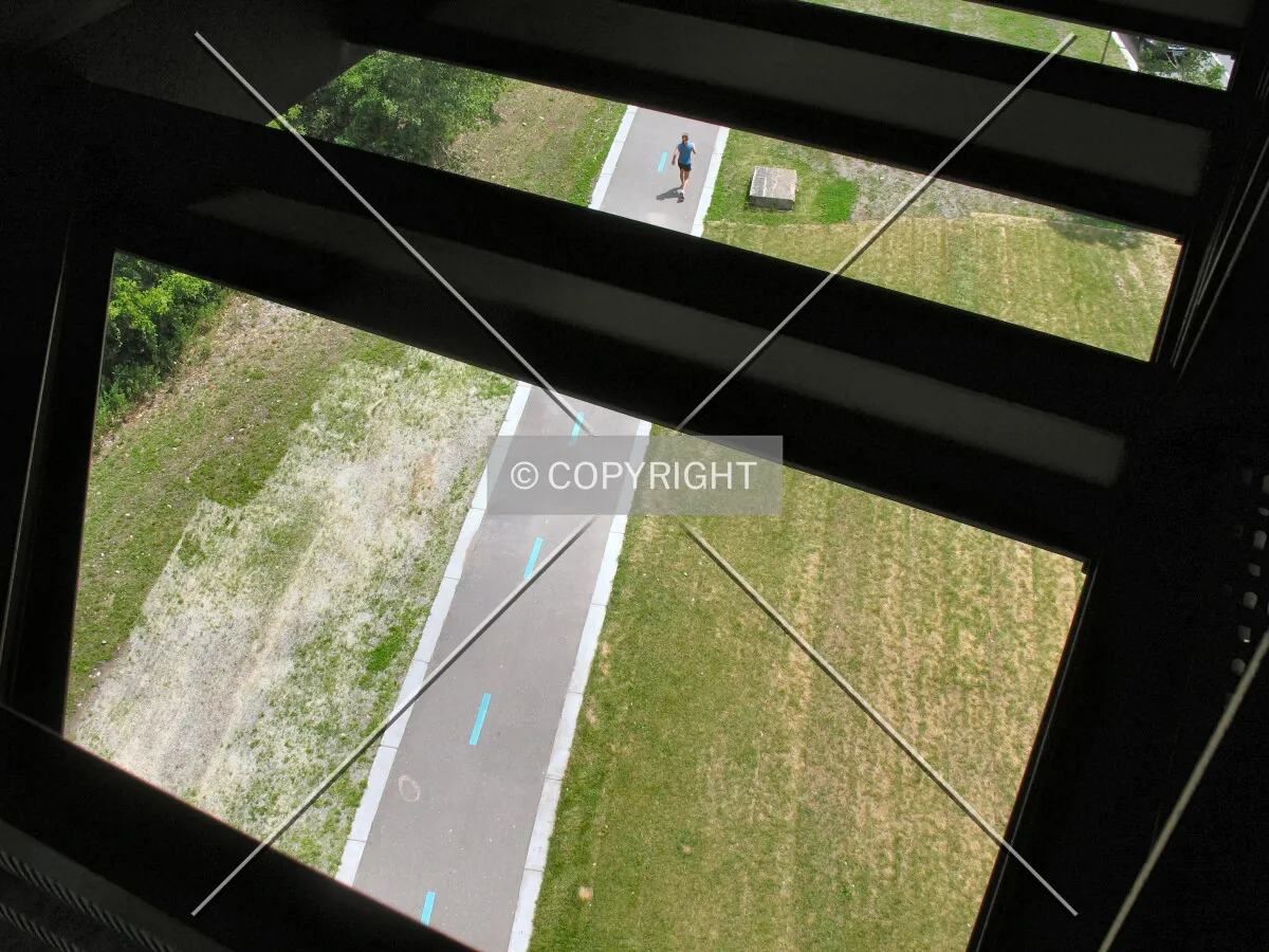 Looking down from below the upper terrace on the Endless Bridge, structural supports contrast with a riverside recreation trail